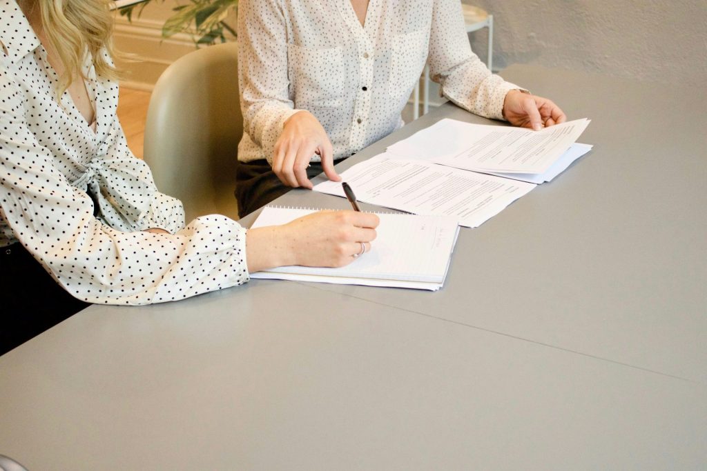 Eine Frau unterschreibt auf weißem Druckerpapier, neben ihr eine Frau, die im Begriff ist, die Dokumente zu berühren.